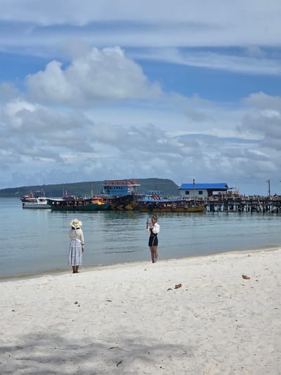 Two women stand on a white sandy beach in Koh Toch Village, Cambodia. The woman on the left faces the water, wearing a straw hat, a white long-sleeved top, and a long blue and white striped skirt. The woman on the right faces the viewer, holding a phone in her left hand, wearing a white jacket with black trim over a black skirt and dark shoes. She appears to be taking a picture. In the background, several boats are moored in the calm water, some of which appear to be tour boats. A wooden pier with buildings on it extends into the water to the right. The sky is blue with scattered white clouds. The water is clear and calm, lapping gently at the shore.