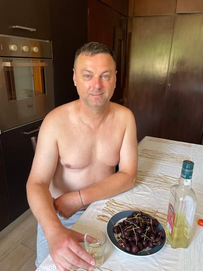 The image shows a topless middle-aged man with blue eyes and short brown hair sitting at a table indoors. He is wearing grey sweatpants. In front of him is a plate of dark cherries, a shot glass with a clear liquid, and a bottle of yellow liquid labeled "TISCAZ MEXICO". A small red tomato is visible to the right of the bottle. The man is looking directly at the camera. The background features a kitchen with dark cabinets and an oven. The table is covered with a white tablecloth featuring a gold linear pattern. The setting appears to be a residential kitchen.