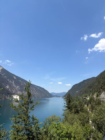 The image shows a scenic view of a mountain lake in Pertisau, Austria. The scene is dominated by the vast blue sky, with a few wispy clouds scattered across it. A faint contrail from an airplane is visible high in the sky. In the foreground, lush green trees and pine branches frame the lower right portion of the image.
The middle ground features a serene, blue lake that stretches across the frame, reflecting the clear sky. The water appears calm, with a few small sailboats visible in the distance, suggesting a leisurely day on the water. Steep, tree-covered mountains rise dramatically from the opposite shore of the lake, their slopes dotted with dense green foliage. The mountains appear rugged and imposing, creating a dramatic backdrop.
The overall impression is one of natural beauty and tranquility. The time of day appears to be daytime, with bright sunlight illuminating the landscape and casting subtle shadows. The weather seems clear and pleasant, ideal for outdoor activities. The location context of Pertisau, Austria, suggests the presence of alpine scenery and a popular tourist destination. No text is visible in the image.