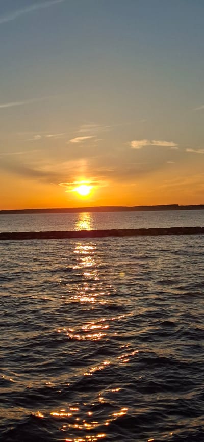 A vertical, close-up shot depicts a body of water under a sunset. The sun, a bright orb reflecting on the water, is located in the upper half of the frame, casting a golden glow across the horizon and the rippling surface of the water. The water is dark, with the reflection of the sun creating a shimmering path. A thin, horizontal line of dark rocks or a structure is visible in the middle ground, separating the sun's reflection from the main body of water. The sky transitions from a pale blue at the top to a warm orange near the horizon, with faint clouds scattered across it. No people or specific landmarks are visible to indicate a precise location within Pärnu linn, Estonia. The scene is natural and serene, capturing the end of the day.