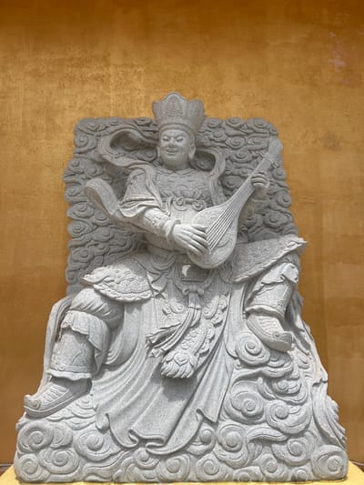 A grey granite statue of a regal figure is centered against a textured golden-yellow wall in Đăk Mar, Vietnam. The figure, depicted seated with a benevolent smile and closed eyes, wears an elaborate crown and intricate robes with armor-like elements. It is actively engaged in playing a stringed, lute-like instrument with both hands. The figure is carved upon and surrounded by stylized cloud patterns that form its base and backdrop within the overall stone carving. The stone exhibits a speckled, light grey texture.