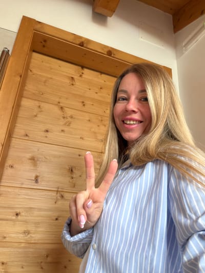 A blonde woman with blue eyes and fair skin smiles at the camera, holding up two fingers in a peace sign. She is wearing a light blue and white striped collared shirt. Her fingernails are painted with French manicures. The background is a wooden wall with horizontal planks and visible knots. The scene is likely indoors, possibly in a residential or hospitality setting in Sauris / Zahre, Italy, given the rustic wooden interior often found in the region.