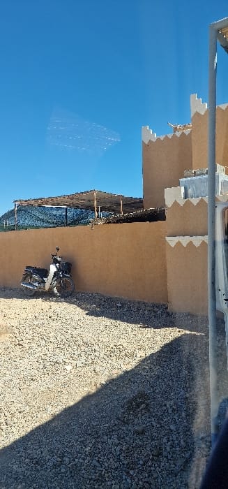 The image displays an outdoor scene in Ferkla El Oulia, Morocco, under a clear blue sky. A light-colored gravel ground occupies the foreground and mid-ground, bisected by a sharp, diagonal shadow extending from the lower right towards the upper left. A white and black motorcycle, equipped with a front basket, is parked leaning against a smooth, tan-colored wall situated on the left side of the frame. This wall extends horizontally, behind which a shaded structure with a light-colored fabric or tarp roof supported by wooden posts is visible. Dark netting or fencing is present beneath this shaded structure, with a distant, low mountain or hill range beyond it in the background, partially covered by what appears to be green mesh or agricultural netting. To the right, a section of a building featuring traditional Moroccan architecture is visible, constructed of tan-colored material with distinct white, crenellated coping along its upper edges. No individuals are present. Reflections on the left side of the frame suggest the image was captured through a transparent barrier.