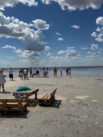 The image depicts a sunny day at a salt lake beach in Бурсоль, Russia. The sky is a vibrant blue, dotted with fluffy white clouds. In the foreground, there are several makeshift wooden furniture pieces constructed from pallets, serving as benches and a table. A teal towel and a water bottle are visible on the table. The ground is a light-colored, dry salt crust with scattered debris.
In the mid-ground and background, numerous people are enjoying the water and the beach. They are dressed in casual summer attire, with some wading in the shallow, reflective water, while others stand on the shore. The lake stretches out to the horizon, appearing calm and shallow. The overall scene conveys a sense of leisure and recreation at a unique natural destination.