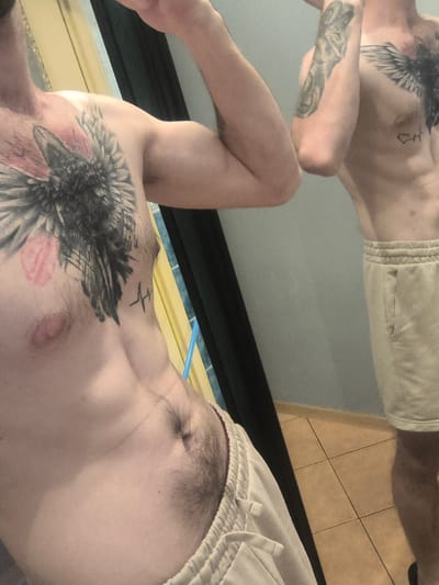 The image shows a close-up, distorted reflection of a person's torso and arms. The person has a prominent tattoo of a bird of prey on their chest and another tattoo on their forearm. They are wearing light-colored sweatpants. The background suggests an indoor setting with tiled flooring visible and a wall in a muted color. The lighting is soft, and the overall impression is casual, possibly taken in a bathroom or bedroom due to the reflective surface. There is no explicit indication of the time of day or weather. No visible text is present in the image. The location context of Grójec, Poland is noted but not visually evident within the image itself.