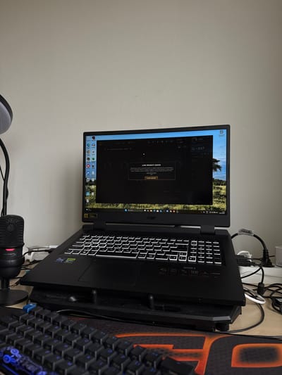 The image shows a laptop computer sitting on a desk. The screen displays a video game interface with the text "LOW PRIORITY QUEUE" and "LEAVE QUEUE," suggesting the user is in a waiting state within the game. The laptop appears to be an Acer Nitro 5 model, as indicated by the branding on the keyboard area. Adjacent to the laptop is a gaming keyboard with blue backlighting, and a HyperX microphone is also present. The location of this scene is not discernible within the city of Jönköping, Sweden.