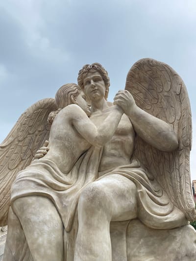 The image depicts a stone sculpture of two winged figures embracing. The figures are rendered in a classical style, with draped clothing and detailed musculature. The larger figure, presumably male, has a muscular torso and is seated, with its arms wrapped around the smaller figure. The smaller figure, leaning into the larger one, has its head resting on the larger figure's chest and its arms around its neck. Both figures have wings, intricately carved to resemble feathers, which extend upwards and outwards. The sculpture is a light, creamy beige color and appears to be made of stone. The background is a pale, overcast sky. This sculpture is located in Nghĩa Trụ Commune, Vietnam, specifically in front of a temple, likely the Bát Tràng Temple.