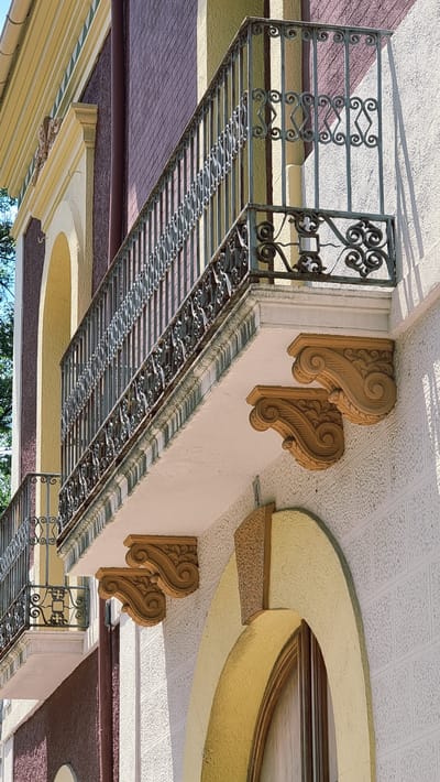 The image captures a detailed view of a building facade in Olot, Spain, focusing on a decorative balcony and its supporting elements. The building features a textured white exterior with textured maroon and yellow sections. A wrought iron balcony railing, intricate with scrolling patterns and vertical bars, extends across the frame. Beneath the balcony, ornate, curved wooden brackets with carved floral and scroll motifs are prominently displayed, supporting the balcony's edge. Another smaller balcony, with a similar railing, is visible on a lower level. An arched window with a wooden frame is partially visible on the right side of the image, framed by yellow molding. The lighting suggests a sunny day, casting shadows on the building's surface. The composition emphasizes architectural details and the interplay of light and shadow on the building's textured surfaces.