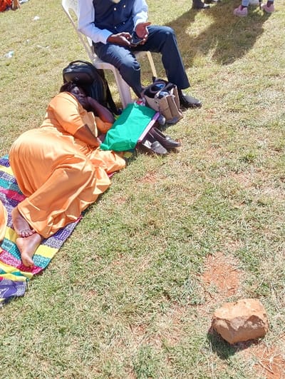This image depicts a relaxed outdoor scene in Kimumu ward, Kenya, under bright daylight. In the foreground, a woman wearing a long, flowing orange dress is lying on her side on a colorful patterned blanket. She appears to be resting or sleeping, with her eyes closed or squinting and her bare feet visible on the blanket.
Adjacent to her, a man is seated on a white plastic chair. He is dressed in a white shirt, a dark blue vest, and dark trousers, and is looking down at a smartphone he holds in his hands. A black backpack rests on the ground behind his chair. Several pairs of shoes, including dark loafers and boots, are casually placed on the grass near his feet, alongside a bright green bag. The legs of another person, possibly a child, wearing pink shoes, are partially visible in the upper background.
The setting is an open grassy area, with patches of green and dry grass visible across the ground. The strong, clear shadows indicate a sunny day, likely during mid-morning or afternoon. A prominent, irregularly shaped light brown rock sits on the grass in the lower right portion of the frame. The overall atmosphere suggests an informal gathering or a moment of leisure outdoors, characteristic of a community event or family outing. There is no visible text in the image.