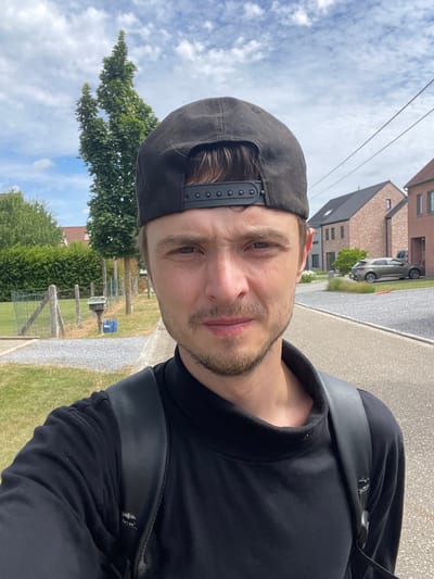 A young man, wearing a black t-shirt and a black baseball cap, stands outdoors, taking a selfie. He has a light beard and stubble. A black backpack is visible on his shoulders. In the background, to the left, a green hedge lines a fence, with a small metal mailbox attached to one of the fence posts. A blue crate sits on the grass next to the mailbox. Further back, there are trees and houses with red-tiled roofs. To the right of the man, a paved road leads past more houses and a parked car. The sky is partly cloudy. The setting appears to be a residential street in Diepenbeek, Belgium.