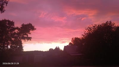 This is a photograph of a sunset over a rural landscape, likely on the outskirts of Луза, Russia. The sky is filled with vibrant pink and purple clouds, indicating either a sunrise or sunset. In the foreground, silhouetted trees frame the scene on the left and right. Several low-lying structures, possibly outbuildings or sheds, are visible on the left side of the image, partially obscured by vegetation. A house with a dark, sloped roof is situated in the middle ground on the right. A line of trees forms a dark silhouette against the horizon in the distance. The lower-left corner of the image contains a timestamp: "2026/06/28 21:29". There are no people or animals visible in the image.