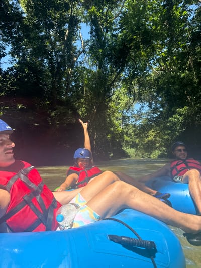 Three individuals are engaged in river tubing on a sunlit river, identifiable as occurring in Woods Town, Jamaica. The scene is dominated by dense, green tropical foliage lining both banks, forming a canopy overhead with patches of clear blue sky visible. The water is a greenish-brown with visible ripples.

In the foreground, partially visible on the left, is a male subject wearing a red life vest, a blue helmet, and patterned pastel shorts. A clear plastic water bottle is secured between his legs and the blue inflatable tube he occupies. His face, partially obscured, is directed left.

In the center, a female subject smiles, her left arm raised high with fingers spread. She wears a blue helmet with a white "NRS" logo, a red life vest, and patterned shorts. She is seated in a blue inflatable tube.

On the right, a second male subject is visible, wearing a red life vest, dark helmet or sunglasses, and dark shorts. He is seated in a separate blue inflatable tube, his legs extended, and dark water shoes are visible. All three individuals appear to be floating downstream.