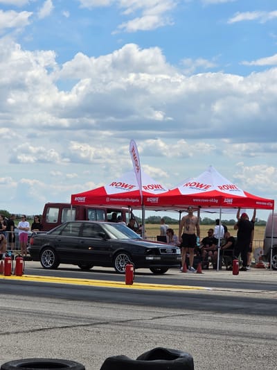 The image captures a scene at a drag racing event held in Akatsievo, Bulgaria. A black Audi sedan is positioned on the asphalt track, flanked by yellow lines and tire marks. In the background, several white and red tents with "ROWE MOTOR OIL" branding are set up, suggesting sponsorship. A crowd of people, dressed in casual attire, are visible near the tents, some observing the event. A man with tattoos on his chest stands prominently near the tents, wearing black shorts and a bandana. The environment is outdoors under a cloudy sky, with indications of open fields beyond the track. Fire extinguishers are placed near the car and tents, indicating safety precautions.