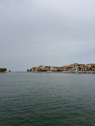 The image displays a waterfront scene in Higüey, Dominican Republic. The composition is divided vertically, with the left side dominated by a hazy, overcast sky and the right side featuring a body of water alongside a row of buildings. These buildings appear to be apartment or hotel complexes, characterized by their tan and beige facades, arched windows, and multiple stories. Palm trees are visible interspersed among the structures, suggesting a tropical or subtropical climate.
The water is a dark, textured gray, with visible ripples indicating a slight choppiness on the surface. Several boats are docked along the waterfront, the most prominent being a sleek, white vessel with multiple levels. A marina or private dock area is apparent, providing moorings for these boats. In the distance, the shoreline recedes, with more buildings and greenery visible, hinting at a developed coastal area.
The sky is a uniform, muted gray, suggesting either early morning, late afternoon, or an overcast day. There are no discernible people in the image, and the focus is on the architecture, the water, and the natural elements. The overall impression is one of a tranquil, possibly residential or resort-style, waterfront environment. No text is visible in the image.