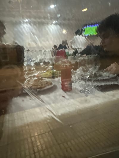 The distorted image appears to show a reflection or view through a rain-covered surface. Colors like red, yellow, green, and blue are visible. Indistinct shapes suggest buildings, objects, or people. The surrounding area consists of various shades of white and grey. The overall impression is a blurred and unclear street scene or interior space, obscured by a water-covered glass or similar surface.