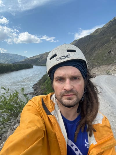 A close-up selfie depicts a Caucasian male with brown dreadlocks, wearing a white climbing helmet with a dark blue headband, a blue t-shirt with a white design, and an orange jacket. The man has dirt on his face. The background shows a landscape with mountains, a cloudy sky, and a river. The location is identified as Edigan, Russia.