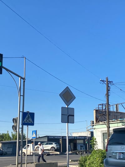 This media file depicts a street scene in Төмөнкү Ала-Арча, Kyrgyzstan, under a clear blue sky, suggesting a sunny day. The image is oriented vertically, with the sky occupying the upper portion.

Prominently featured are traffic signs. There's a diamond-shaped sign with a white interior and a gray border, likely indicating a priority road or main road. Below it, a blue square sign with a white pedestrian symbol indicates a pedestrian crossing. Another smaller blue sign with a white pedestrian symbol and arrows suggests a designated pedestrian path. Traffic lights are visible, with at least one green light illuminated, indicating that traffic may proceed.

In the background, buildings line the street. One building has a facade with text that includes "ПЛАСТИКОВЫЕ ОКНА" and "ВИТРАЖИ", indicating it is a business selling plastic windows and stained glass. Several vehicles are present, including a white Lexus SUV with a license plate that appears to be from Kyrgyzstan and a white utility vehicle.

A person, dressed in a white shirt and dark pants, is walking across the street, likely near the pedestrian crossing indicated by the signage. The overall scene suggests a normal day in a town or city with active traffic and commerce. The time of day appears to be daytime, given the bright sunlight and clear sky. There are no overt emotional cues, but the presence of people and vehicles suggests a functioning urban environment. Overhead power lines are also visible, crisscrossing the sky.