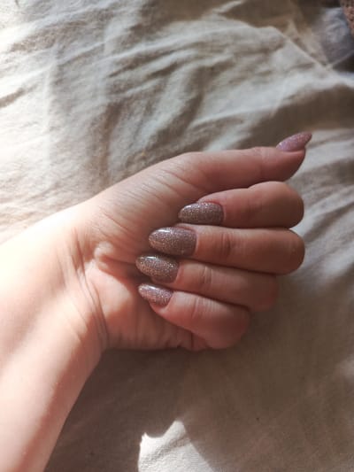 The media file is a close-up shot of a person's left hand, with a focus on their fingernails. The hand is resting on a light-colored, textured fabric, possibly a blanket or sheet, which is creased and shows shadows from overhead lighting. The fingernails are uniformly shaped and painted with a shimmery, rose-gold or light brown glitter polish. The lighting suggests it is daytime, with sunlight casting strong shadows on the fabric. The overall scene is intimate and personal, highlighting the detail of the manicured nails. There are no visible text elements in the image. The location context provided is Salavat, Russia, though this cannot be deduced from the visual content alone.