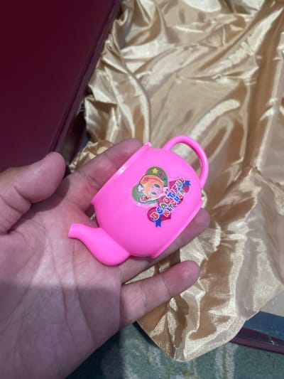 A hand holds a small, bright pink plastic teapot. The teapot has a sticker on it with a cartoon character wearing a hat and the words "BEAUTIFUL KITCHEN". The teapot is resting on a surface covered with a shimmering, golden fabric. Behind the fabric, a dark red box is partially visible. The scene appears to be indoors, possibly in a room in Kara-Oi, Kyrgyzstan.