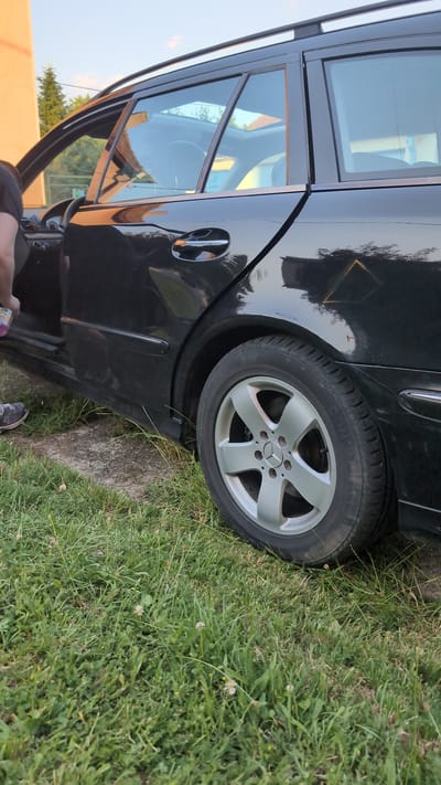 The image shows the side of a black Mercedes-Benz station wagon. The rear passenger door is ajar, revealing part of the interior and the car's window. The car is parked on a grassy area next to a concrete path. A person's legs and feet, wearing sneakers and dark pants, are visible in the lower right corner of the image. The person is holding a colorful object in their left hand. The wheel of the car has a silver five-spoke rim with the Mercedes-Benz logo in the center. The time of day appears to be late afternoon or early evening, with soft sunlight casting shadows and highlighting the car's glossy finish. The grass is green and appears to be well-maintained. The scene is outdoors, likely in a residential area or a park, given the presence of grass and a concrete path. There is no visible text in the image.