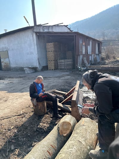 In Dobrodan, Bulgaria, an outdoor industrial area features two adult males in a working environment. The scene is illuminated by direct sunlight, casting distinct shadows.
In the mid-ground, a light-colored concrete building with a corrugated metal roof stands to the left, from which a tall, slender metal chimney ascends. An open bay on this building reveals numerous orderly stacks of light-colored wooden pallets. To the right of the concrete structure is an older, distressed building clad in rusted corrugated metal panels, also with a partially damaged roof. The background features a tree-covered mountain range beneath a clear sky. The ground throughout the visible area is uneven dirt and mud, with scattered wood chips and debris.
Two large, debarked logs lie horizontally across the foreground. The left log displays a clean circular cut end, while the right log exhibits several vertical gouge marks on its surface.
**Subject 1 (Left):** An adult male, bald, is seated on a vertical log section. He wears a blue t-shirt, an open dark zippered hoodie, dark trousers, and dark work boots. He holds a black smartphone in both hands and is focused on its screen, angled slightly to his right.
**Subject 2 (Right):** An adult male, bald, wears a dark hooded sweatshirt with the hood pulled up over his head, dark trousers, and dark work boots equipped with a blue circular fastening mechanism. A red bracelet is visible on his left wrist. He is positioned in a crouch or low-seated stance, partially obscured by the foreground logs, and holds a black smartphone in both hands, actively observing its screen, angled slightly to his left.
Behind Subject 1, a reddish-brown metal frame structure, possibly a piece of equipment or support, is visible, featuring a heavy metal chain connected to a horizontal plate. Both subjects are independently engaged with their mobile phones; no direct interaction between them is observed.
