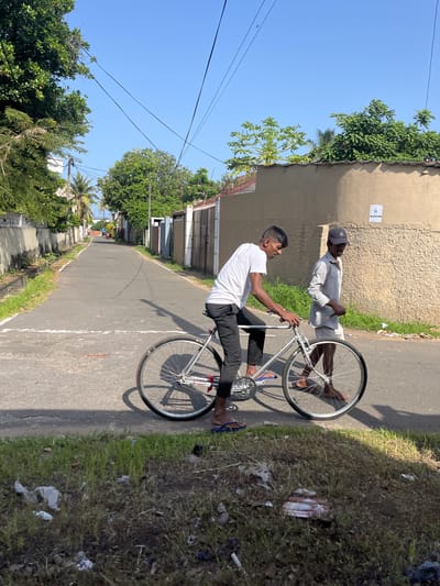 The image shows two men on a street in Moratuwa, Sri Lanka. The man in the foreground is wearing a white t-shirt and black pants and is riding a bicycle. He is wearing blue flip-flops. The man behind him is wearing a light gray shirt and shorts and a dark baseball cap. He is walking and holding a stick in his right hand. The street is paved and has a white line painted across it. There are buildings and trees on both sides of the street. The sky is clear and blue. The scene appears to be a typical street in a residential area.