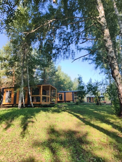 In Катунь, Russia, a cluster of modern, modular cabins with wooden exteriors and large windows is nestled among tall trees. The cabins are elevated on stilts, surrounded by a grassy area with dappled sunlight casting shadows. The sky is clear and blue. The scene appears to be a tranquil, natural setting, possibly a resort or glamping site.