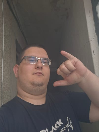 The image displays an adult male, likely in an indoor or semi-sheltered outdoor setting. The subject is overweight, with short dark hair, and wears rectangular glasses with silver-colored frames. A visible stubble covers his chin and jawline, emphasizing a double chin. He is wearing a dark, possibly navy blue or black, crew-neck t-shirt with white text on the lower left side, partially readable as "ACK yourself". The male is looking directly at the camera. His right hand is raised, performing a gesture with the thumb and pinky extended and the three middle fingers curled into the palm.
The background consists of two distinct wall sections. To the subject's left, a light brown or beige wall exhibits a rough, textured surface, possibly stucco or coarse concrete, with a thin white cable or cord running vertically along it. To the subject's right and behind him, a darker, smoother, grey-colored wall is visible. A vertical architectural feature, possibly a door frame or column, separates these two wall surfaces. The darker wall shows some faint marks or discoloration, and a dark, thin crack is present in the upper right portion of the background. There are no discernible visual elements within the image to confirm the specific location as Kran, Bulgaria.