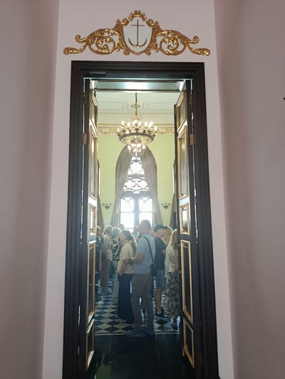 In the city of Kosava, Belarus, an open doorway with dark wooden frames and gold trim reveals a grand hall. The hall features a black and white checkered floor, a large chandelier suspended from the ceiling, and tall, arched windows draped with light-colored curtains. A group of people, appearing to be tourists, stand inside the hall, some facing towards the windows and others looking at each other. Above the doorway, a decorative gold emblem with a white shield and an anchor symbol is affixed to the wall. The scene suggests an interior view of a historical building or museum, possibly the Pushkin Museum in Kosava.
