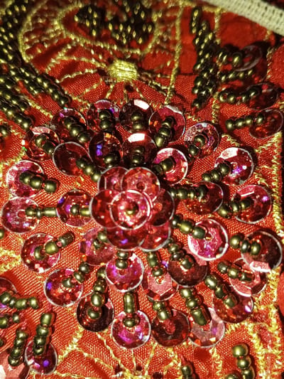 This image presents a forensic close-up of an intricately embellished red textile, located in the city of Keo Kouang, Laos. The primary subject is a densely decorated fabric, possibly silk or satin, characterized by its vibrant red hue and visible weave texture.
The fabric is adorned with numerous circular, flat, translucent red sequins, each approximately 5-7mm in diameter. Many sequins exhibit an iridescent quality, reflecting subtle purple and pink light. Each sequin is secured to the fabric by small, metallic, dark gold-bronze seed beads, typically arranged in a cluster of two to three beads passing through the central hole of the sequin.
A prominent central arrangement features several sequins clustered together to form a flower-like pattern. Radiating outwards from this central cluster, and interspersed across the fabric, are individual sequins and lines of the dark gold-bronze seed beads. These beads are frequently strung in short segments of three to four beads, creating small, elongated decorative elements.
Underneath and around the sequin and bead work, intricate patterns of metallic gold thread embroidery are visible. These embroidered lines form flowing, curvilinear designs that complement the beaded embellishments. The lighting highlights the reflective surfaces of the sequins and beads, creating varying degrees of sheen and shadow. No individuals or actions are depicted in the scene; the focus is exclusively on the textile's detailed ornamentation.