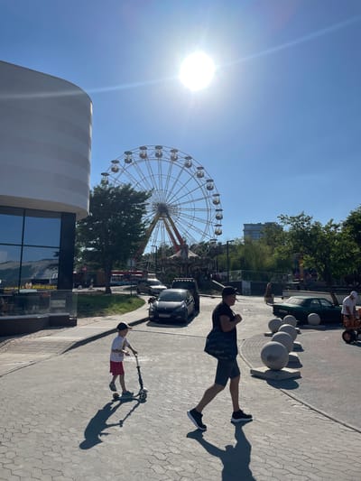 In Merchanskoye, Russia, on a sunny day, a child rides a scooter on a paved area. A man walks past, carrying a bag over his shoulder, his shadow cast long on the ground. In the background, a Ferris wheel stands tall, and several cars are parked along the street. Trees and buildings complete the urban landscape, under a clear blue sky.