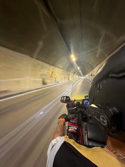 The image is a first-person perspective shot from a motorcycle traveling through a tunnel. The tunnel walls are made of a light-colored material and are illuminated by overhead lights. There are white lines marking lanes on the road. The rider's arm is visible in the foreground, wearing a black glove with an orange band and a tattoo on the forearm. The motorcycle's dashboard is also visible, with a speedometer and other gauges. The scene appears to be in motion, with a slight blur indicating the motorcycle's movement. The location is stated to be in the city of Đurmani, Montenegro, though this specific detail cannot be verified from the visual information alone.