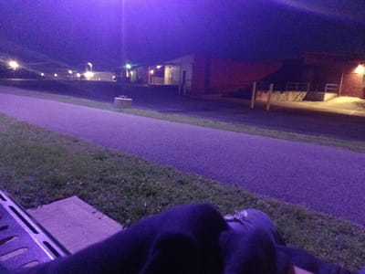 The image depicts a nighttime outdoor scene, heavily saturated with a purple or violet hue across the entire frame. In the immediate foreground, what appears to be a person's lower body, possibly legs and torso, is visible, clad in dark clothing (suggesting a hoodie or similar casual wear). To the left of the person, a metallic, grated structure, possibly a railing or part of a bench, is partially visible.

The midground features a paved path or sidewalk, which stretches diagonally from the left foreground towards the center-right background. Lush green grass borders the path on both sides. To the right of the path, a small, round, light-colored concrete object, possibly a planter or bollard, is situated on the grass.

In the background, several buildings are illuminated by artificial lights, creating bright flares in the dark sky. These structures appear to be commercial or industrial, with some featuring loading docks or service entrances, particularly on the right side where ramps and railings are discernible. Streetlights are also visible, casting their glow on the scene.

The dominant purple color suggests specific artificial lighting conditions or a camera's low-light performance. The time of day is clearly night. No specific activity is evident beyond someone sitting or lying down by the path, observing the surroundings. There is no visible text in the image. The setting appears to be an urban or suburban area, possibly near an industrial park or commercial district in Tahlequah, United States of America.