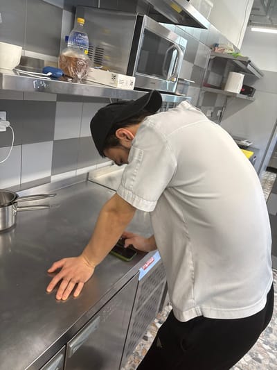 In a commercial kitchen located in Kirkkolakhti, Russia, a male individual is positioned leaning over a stainless steel countertop. He is observed from the upper back and right side, wearing a light-colored, short-sleeved work shirt or chef's jacket, a black baseball cap worn backward, and dark athletic pants. His head is bowed, directed towards the countertop. His left hand is spread flat on the stainless steel surface, while his right hand holds a smartphone with a green case, which is also resting on the countertop directly in front of him. To the left of the individual, on the countertop, rests a small stainless steel pot, possibly nested with another smaller pot.
The immediate background features a tiled wall with a grey and white checkerboard pattern transitioning to plain white above. A stainless steel shelf is mounted above the countertop. On this shelf, from left to right, are a white ceramic mug, a plastic bottle with a blue cap, a clear plastic bottle containing a brown liquid, encased in a plastic bag, a small blue electronic device, and a white box labeled "3F". Above the shelf, a silver Samsung microwave oven is visible, mounted beneath a metallic range hood. Further to the right, an additional stainless steel shelf holds a roll of paper towels and other obscured items. The base units beneath the countertop are stainless steel, featuring door handles and ventilation grates. The floor, partially visible at the bottom right, is tiled with a light, patterned design. A white power outlet with a connected white cord is present on the wall to the far left.