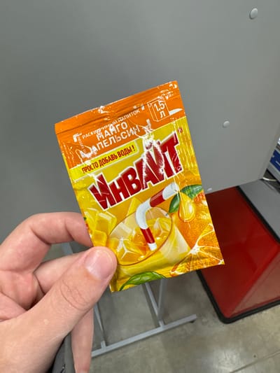 A hand holds an "Invayt" brand instant drink mix packet in Petrovskaya, Russia. The packet has an orange flavor and features images of oranges and candy cane-striped straws. The background includes a light gray surface and a glimpse of a red object with a black outline. The hand appears to be holding the packet for display.