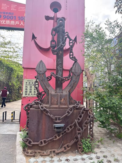 The image shows an anchor-themed display in Dadong District, China. A large red sign with a painted black anchor stands beside a weathered metal structure adorned with a rusty anchor and chains. A sign with Chinese characters indicates that it is a pedestrian area and prohibits motorized vehicles. A person is walking away in the background. The scene is a public space, with trees and sidewalk visible.