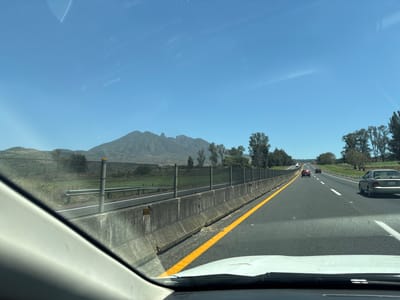 The photograph was taken from the interior of a vehicle traveling on a highway near San Cayetano, Mexico. The mountain, Volcán San Juan, is visible in the distance. A concrete barrier and a chain-link fence run along the left side of the highway. There are multiple vehicles on the road. The sky is clear and blue.