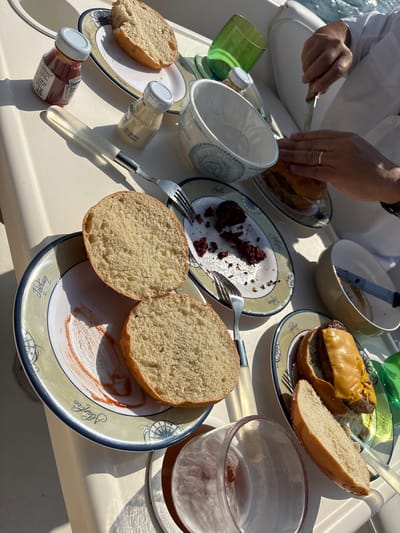 This is a close-up, high-angle, eye-level shot of a picnic lunch on a boat in Callao, Peru. The scene depicts a partially eaten meal of hamburgers and buns on plates. There are two small bottles of condiments, one ketchup and one mayonnaise, and a set of cutlery. In the background, two green glasses and a white bowl are visible, along with the hands of a person wearing a white shirt and a ring, who is cutting into a hamburger. The sunlight casts shadows across the table, indicating it is daytime. The overall environment is that of a casual outdoor meal on a boat.