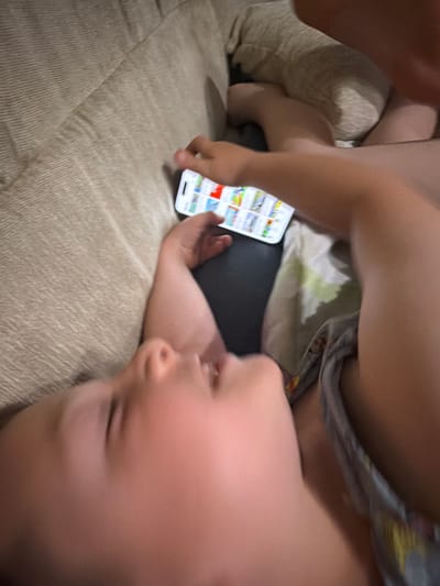 A high-angle, blurry, close-up shot shows a baby lying on a couch, holding a smartphone. The baby's face is in the foreground, out of focus. The smartphone displays a grid of colorful icons, suggesting it is an app or a game. A person's hand is visible reaching for the phone. The couch appears to be made of a beige, textured fabric. The scene is indoors, with soft lighting. The location of Zmeitsa, Bulgaria, is not discernible from the image.