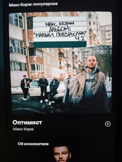 The image shows a screen displaying a music artist's page. At the top, the title "Макс Корж: популярное" is visible, which translates to "Max Korzh: Popular". Below this is a square album cover featuring a photograph. The photograph depicts four men standing and sitting near a white car in an urban environment. Behind them are multi-story apartment buildings with visible balconies. A handwritten sign is superimposed on the album cover, reading "МАКС КОРЖ АЛЬБОМ; "МАЛЫЙ ПОВЗРОСЛЕЛ" 4.2", which translates to "MAX KORZH ALBUM; "YOUNG GREW UP" 4.2".
The main subject in the foreground of the photograph is a man wearing a beige trench coat over a grey Nike hoodie. To his left, slightly out of focus, are three other men. One is standing, leaning against the car, wearing a dark jacket and white sneakers. Two others are seated on the car, one in a dark jacket and the other in a lighter jacket.
Below the album cover, text reads "Оптимист" which means "Optimist", and beneath that, "Макс Корж". Further down, "Об исполнителе" translates to "About the artist". A circular icon with a plus sign is also present. The image is a capture of a digital display, possibly a music streaming service. The question specifies location information about Yalangach, Uzbekistan, which is not discernible from the visual content of the image itself, as the scene is a generic urban setting that could be located in many places.
