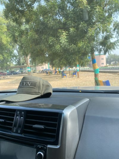 The image depicts an outdoor scene in Yan Dala, Nigeria, viewed from the interior of a vehicle. In the immediate foreground, an olive-green baseball cap featuring a black rectangular patch with the text "ALYN" and horizontal line detailing is positioned on a dark gray, textured vehicle dashboard. Below the cap, central air conditioning vents and a portion of the vehicle's audio system with a control knob are visible.

Beyond the windshield, a park-like area is observed under clear daylight. Numerous trees with dense green foliage are present, several of which have their trunks painted with horizontal bands of vibrant colors, including green, blue, yellow, and orange. Scattered across the dry, light-colored ground are multiple brightly colored plastic chairs, primarily blue and yellow, some accompanied by small tables, indicating an outdoor seating arrangement. To the left, under a shaded structure, several parked vehicles, including cars and motorcycles, are visible. In the background, a multi-story building with light pink or peach-colored walls and horizontal window bands stands prominently. A paved road or pathway with a curb is discernible curving towards the right in the mid-distance. No individuals are visible in the scene.