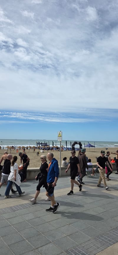 The image shows a group of people walking along a paved walkway adjacent to a beach in Benicàssim, Spain. The sky is visible with a mix of clouds and blue sky. The beach is crowded with various objects and people, including windbreakers. A yellow and black warning sign is visible near the walkway. The walkway is made of gray tiles. People are dressed in a variety of casual clothing, including athletic wear, and are walking in the same direction along the walkway.
