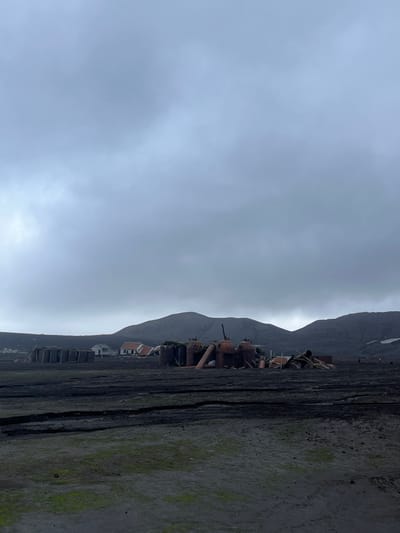 A desolate scene unfolds beneath a gray, overcast sky. Rugged mountains rise in the distance, their peaks disappearing into the clouds. In the foreground, decaying industrial machinery stands on what appears to be a wooden pier or dock. Buildings line the shore, hinting at a once-thriving settlement. The pier itself is cracked and shows signs of wear, with moss growing in the crevices. The somber palette and decaying structures create a sense of abandonment and isolation.