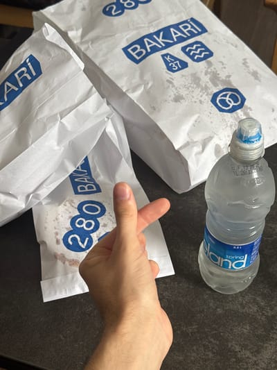 The image shows three white paper bags with blue "BAKARI 280" branding in a stacked arrangement, alongside a 0.5L bottle of "Icland Spring" water, and a partially visible hand making a finger gesture (pinched fingers). The scene suggests food purchase from Bakari 280, a bakery chain located in Reykjavik, Iceland. The setting appears to be an indoor surface. The bottle of water has condensation. The bags have slight staining, possibly from contents.