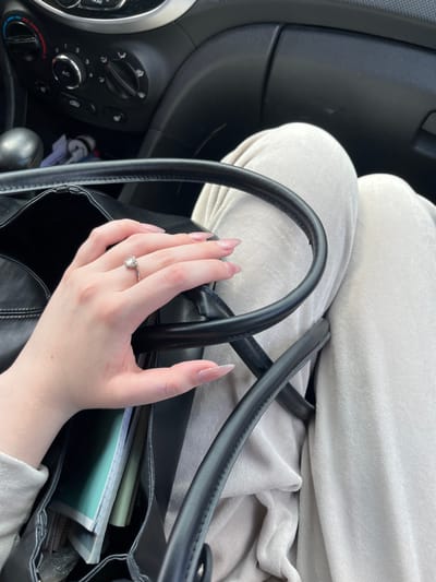 The image shows a close-up of a person's hand with a diamond ring on their finger, resting on the strap of a black handbag. The handbag contains several items, including notebooks. The person is wearing a light-colored, possibly velvet, outfit. The background suggests the interior of a car, with parts of the dashboard and air conditioning controls visible. The scene appears to be within the city of Novaya Otradovka, Russia.