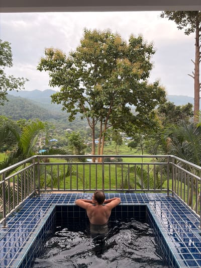 A person is shown from behind, relaxing in a tiled plunge pool on a balcony. The pool is surrounded by a metal railing and overlooks a lush, green valley with hills and trees. In the distance, a small village with houses can be seen nestled within the landscape. The sky is overcast, suggesting a temperate climate. The person's back and shoulders are visible as they lean on the edge of the pool, with their arms resting on the rim. The water in the pool is dark and rippled, indicating movement. The surrounding environment is rich with vegetation, including palm trees and a large deciduous tree with flowering branches positioned prominently in the mid-ground. The scene evokes a sense of tranquility and immersion in nature.