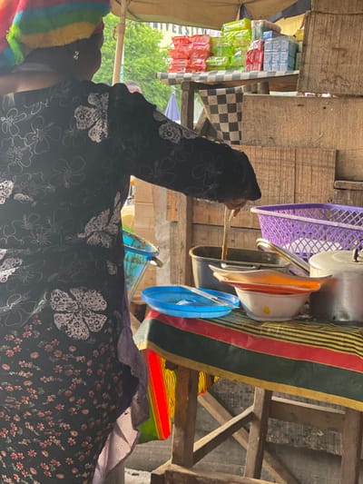 This image captures a vibrant and busy scene, likely within an outdoor market stall or informal shop, viewed from a slightly low angle. The image is rotated 90 degrees counter-clockwise; when viewed upright, it shows the following:
In the foreground, on the left side, a person is visible from the torso upwards, facing towards the right side of the frame. They are wearing a long-sleeved garment, possibly a dress or top, that is black with prominent white floral patterns. A colorful, rainbow-striped (red, orange, yellow, green, blue, purple) fuzzy hat or head covering is worn on their head, obscuring part of their face. Their left hand is raised, holding a thin, light-colored stick-like object, possibly for stirring, pointing, or handing something over. Part of another garment or the same person's layered clothing is visible on the far right, featuring a pattern of smaller, reddish-brown flowers on a dark background, with a checkered purple and white fabric at the neck/collar area.
The background reveals a rustic setup constructed from wooden planks and beams, forming shelves or a display structure. On the left side, various packaged goods are stacked, appearing to be small sachets or packets of products. Some white packets are clearly visible with the text "KLEENO", suggesting tissues, wipes, or a similar household item. Other packets are bright green and red. A black and white checkered cloth is draped over part of the wooden structure near these goods.
On the upper right side of the frame, a variety of kitchen or household items are displayed or stored. These include a purple plastic laundry basket, several metal pots or pans, and various plastic bowls and lids in colors such as white, orange, and blue. These items appear to be hanging or resting on wooden supports. Beneath these hanging items, a large piece of striped fabric with red, green, yellow, and black bands is draped over another wooden structure, possibly a mat or blanket.
In the mid-ground, behind the person and the stall structure, there is a glimpse of an outdoor environment with green foliage, indicating trees. A blue umbrella or canopy is also partially visible, along with a banner that has partial text visible, which appears to read "UNIVERSIT" on a blue background. The scene is brightly lit by natural daylight, suggesting it is daytime. The overall impression is one of an active, informal marketplace with various goods for sale.
