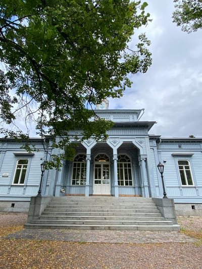 An exterior, eye-level, medium shot captures the facade of a light blue wooden building with a prominent central portico and a grand set of stone stairs leading up to the entrance. The building features white-trimmed windows and decorative architectural elements. Overhead, a large tree with green leaves partially obscures the sky, which is cloudy. The foreground consists of fallen leaves on a cobblestone path, with gravel visible beyond. The scene is set in Sortavala, Russia.
