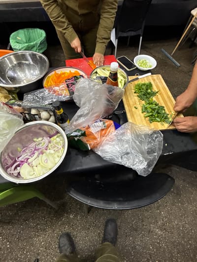 The image shows a scene of food preparation taking place in a kitchen, located in Mazkeret Batya, Israel. A person, dressed in green military-style clothing, is seen cutting green onions with a knife on a wooden cutting board. Another person, wearing brown pants, is not visible in the frame but their hand is seen preparing vegetables, possibly bell peppers, in a black bowl. There are several other bowls containing chopped vegetables, including red onions, and a bag of carrots. A clear plastic bag is laying on the table. The scene is lit with overhead lighting. The table is black. The floor is a gray, rough concrete surface. The background is blurred and shows a chair, a garbage bin, and another person's hand. The image was taken from a low angle, looking down onto the table.