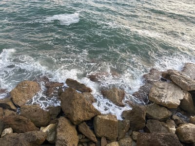 This is a high-angle, eye-level medium shot of waves crashing against a rocky shoreline in the city of Mate Asher Regional Council, Israel. The water is a deep teal color with white foam as waves break against the large, rough, brown and grey boulders that make up the shore. The rocks are varied in size and shape, with some appearing porous and others smooth. The water is clear enough to see the submerged rocks beneath the surface in places. The scene is tranquil and natural. There are no people or animals visible in the image.